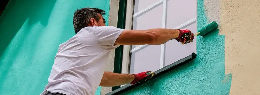 Painter applying teal paint to a home’s exterior wall with roller